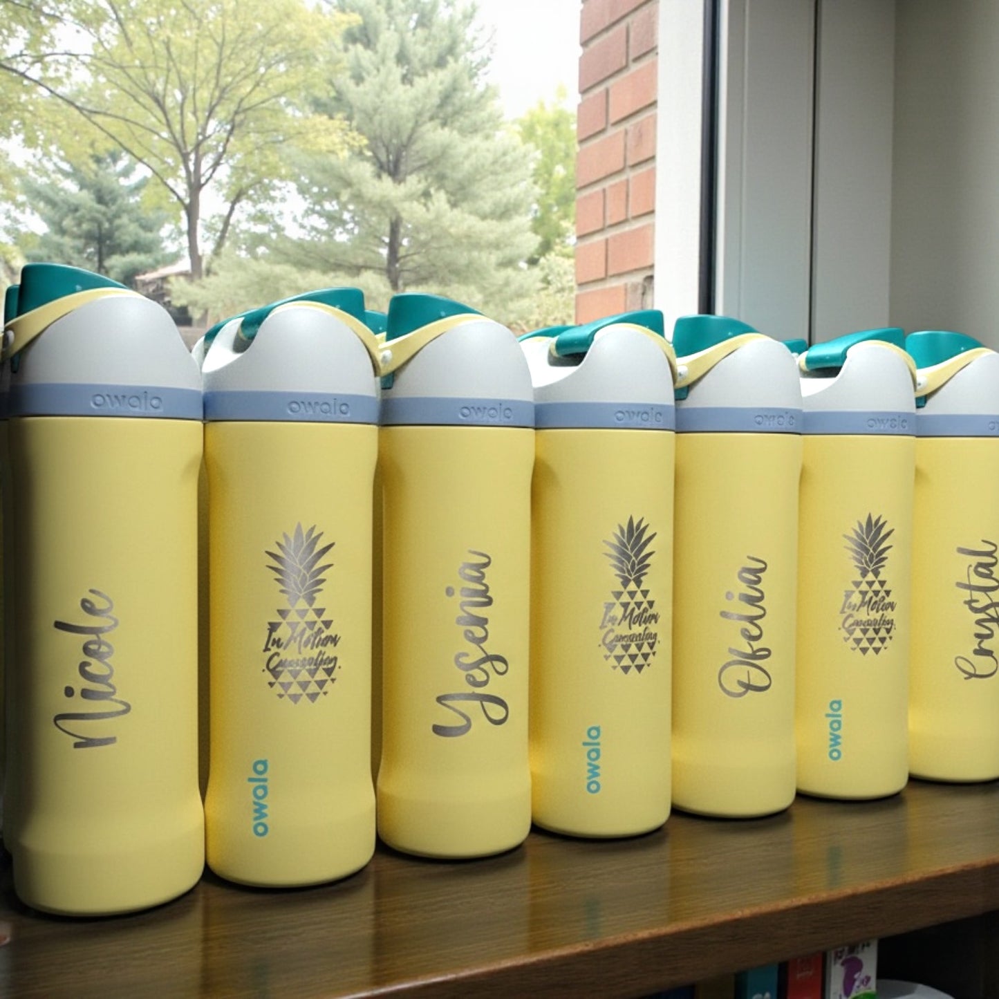 Owala Sunny Daze yellow water bottles with personalized names on a wooden surface near a window.