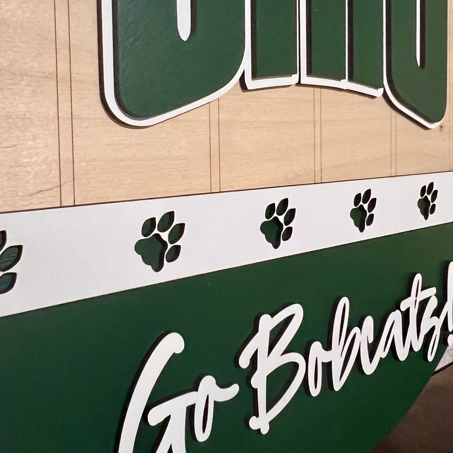 Close-up of Ohio Bobcats door hanger with "Ohio" on top, a band of white with green bobcat paws across the center, and "Go Bobcats!" in white on a green lower half.