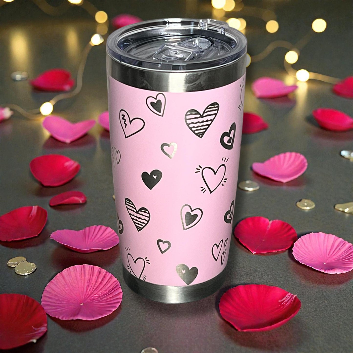 Pink tumbler with heart patterns surrounded by pink petals on a dark surface.