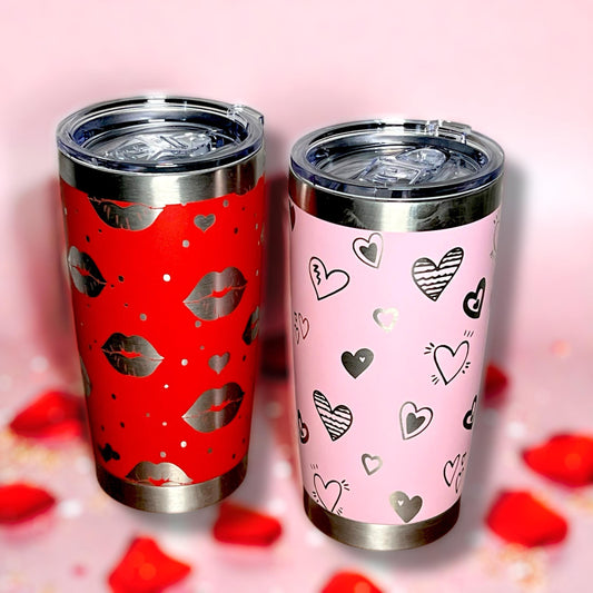 Two tumblers, one red with lip prints and one pink with heart patterns, on a pink background.