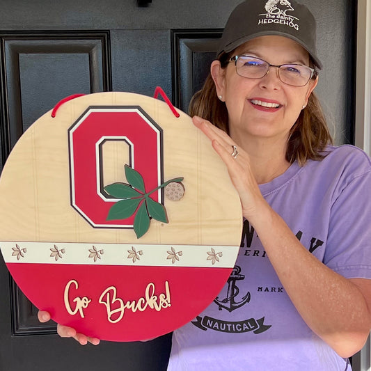 Person holding medium 15” Ohio state buckeyes door hanger with Go Bucks and the block o with buckeye leaf logo.