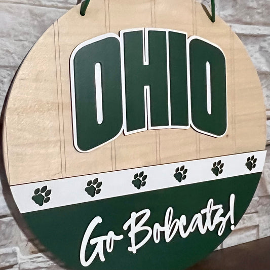 Ohio Bobcats door hanger with "Ohio" on top, a band of white with green bobcat paws across the center, and "Go Bobcats!" in white on a green lower half.