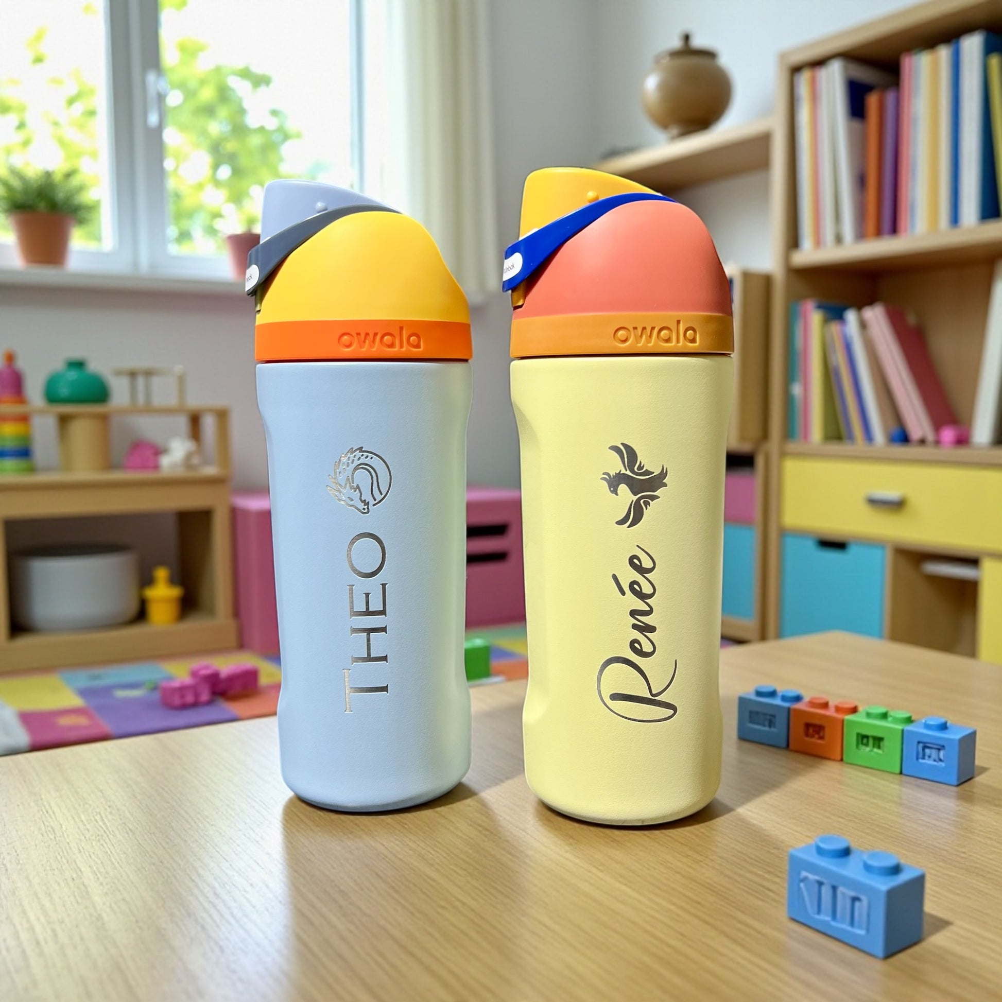 Owala kids. Two colorful water bottles labeled 'Theo' and 'Renée' on a table with a child's play area in the background.