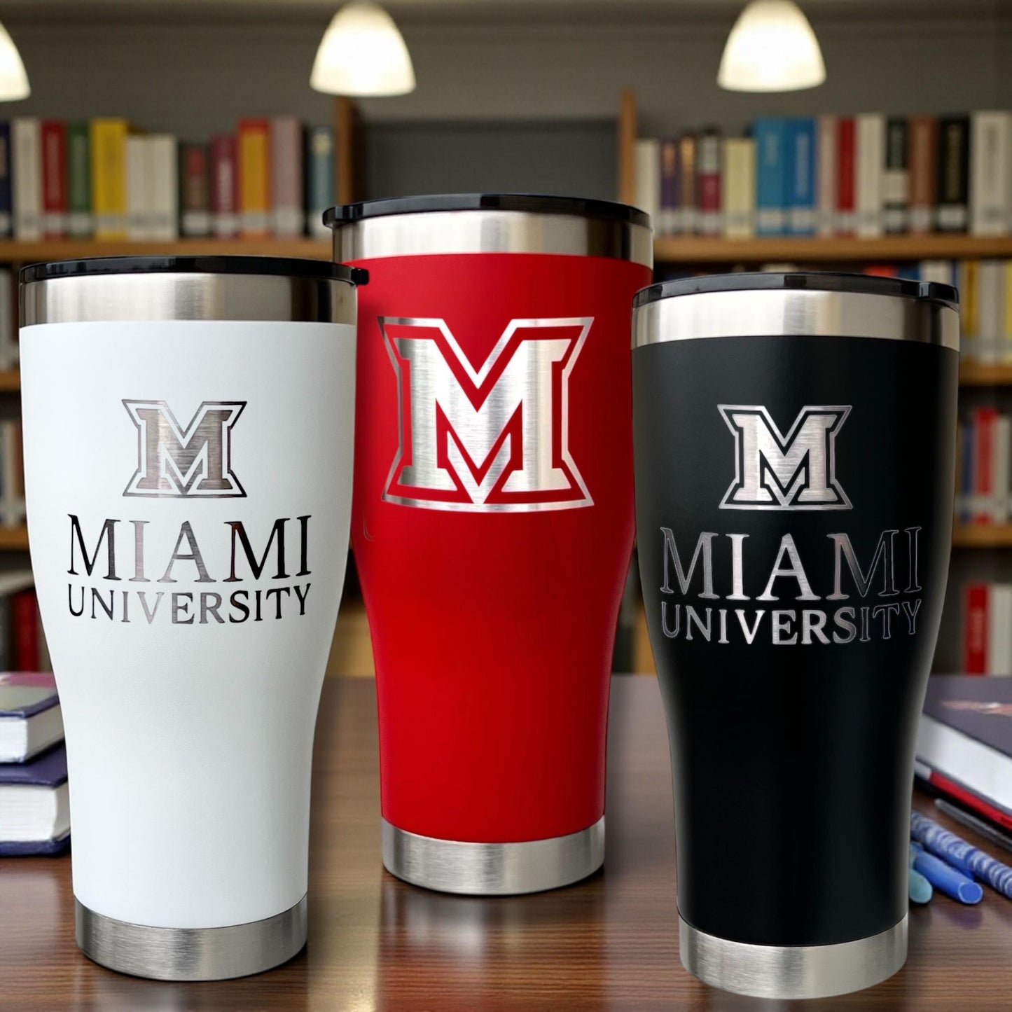Three insulated tumblers with 'Miami University' branding on a table in a library setting.