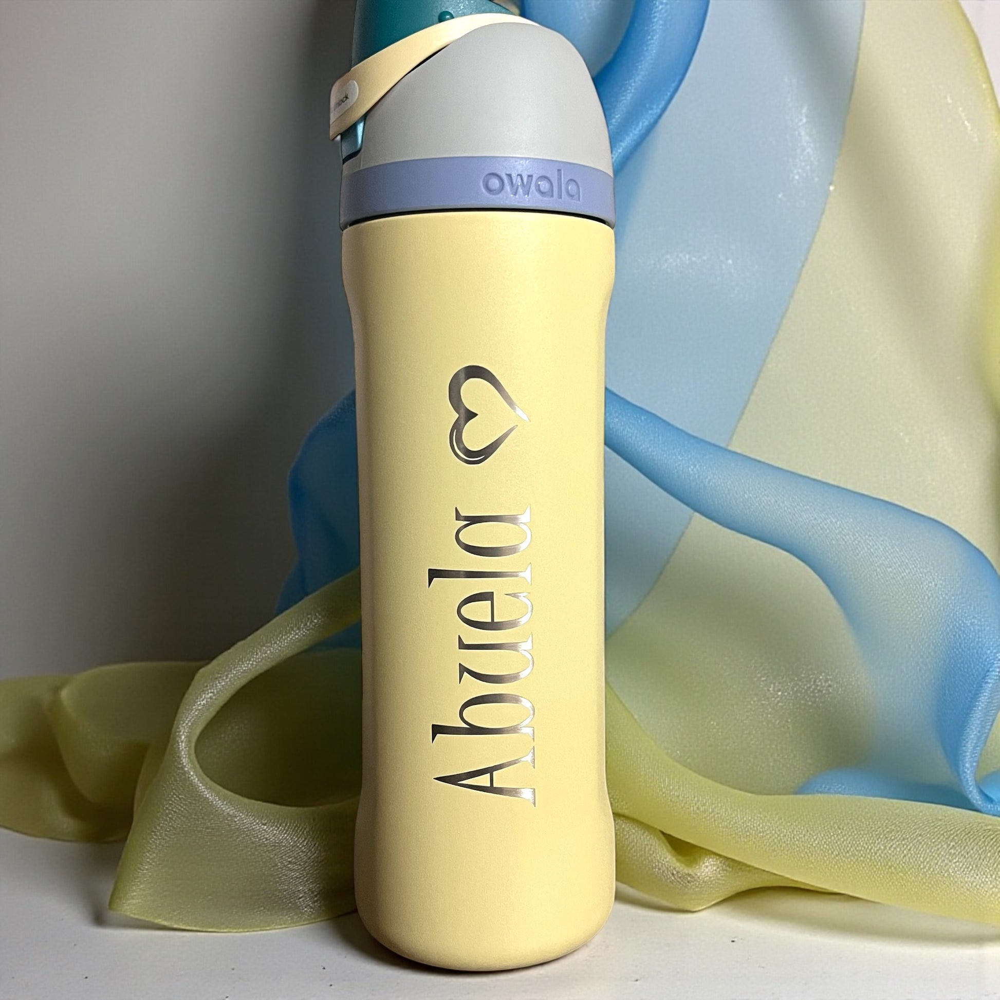 Owala Sunny Daze yellow water bottle with 'Abuela' text and heart logo on a soft fabric background