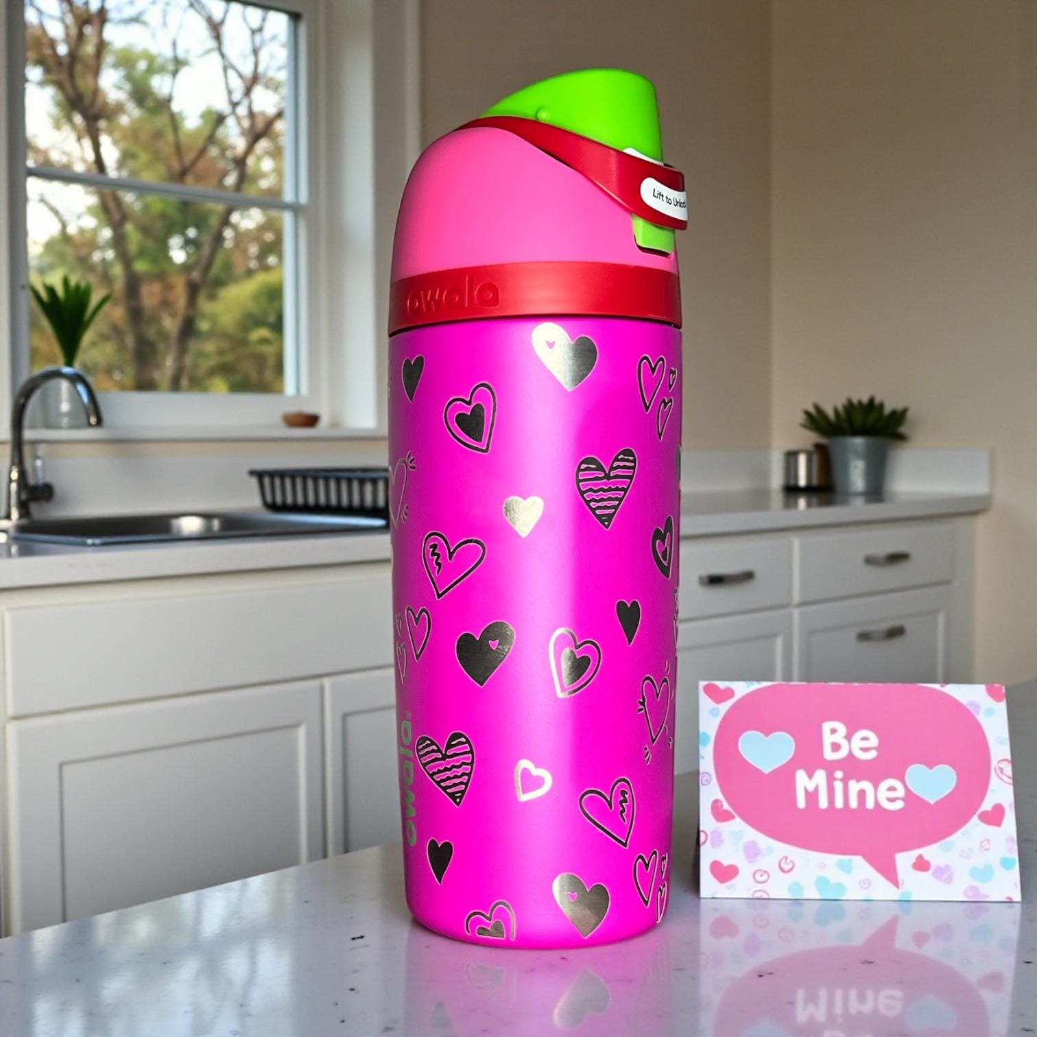 Owala_kids_Valentines_all_the_berries color water bottle with heart patterns on a kitchen counter next to a 'Be Mine' card.
