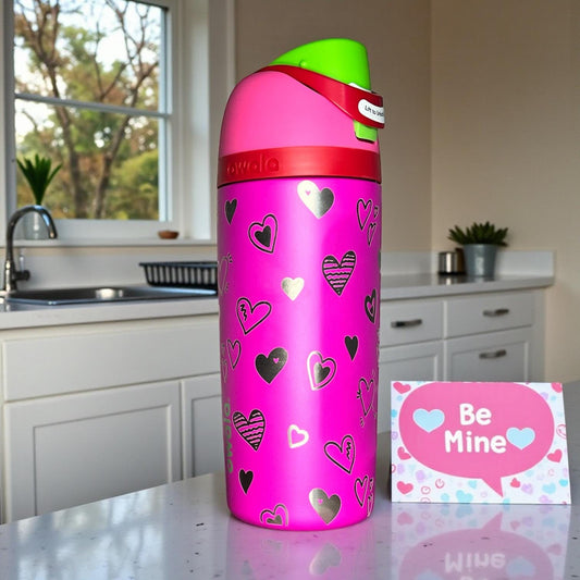 Owala_kids_Valentines_all_the_berries color water bottle with heart patterns on a kitchen counter next to a 'Be Mine' card.